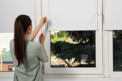 Woman opening white roller blind on window indoors, back view