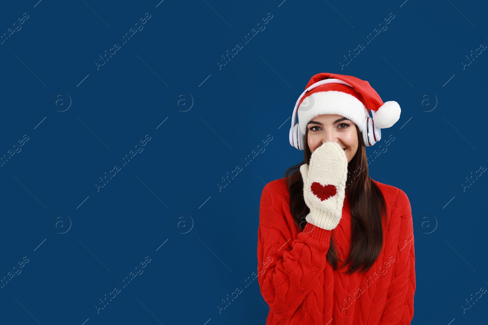 Photo of Young woman in Santa hat listening to Christmas music on dark blue background, space for text