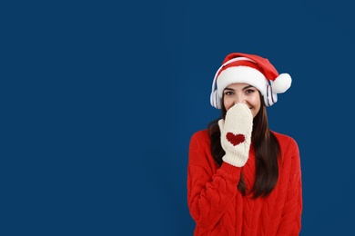 Young woman in Santa hat listening to Christmas music on dark blue background, space for text