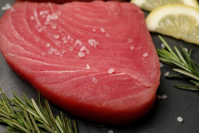 Photo of Raw tuna fillet with rosemary, sea salt and lemon slices on black table, closeup