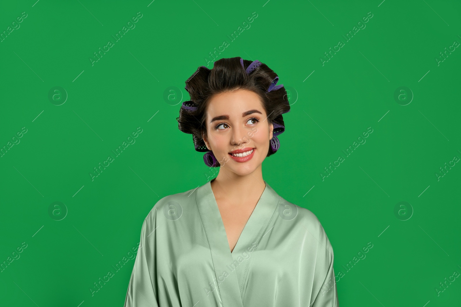 Photo of Happy young woman in silk bathrobe with hair curlers on green background