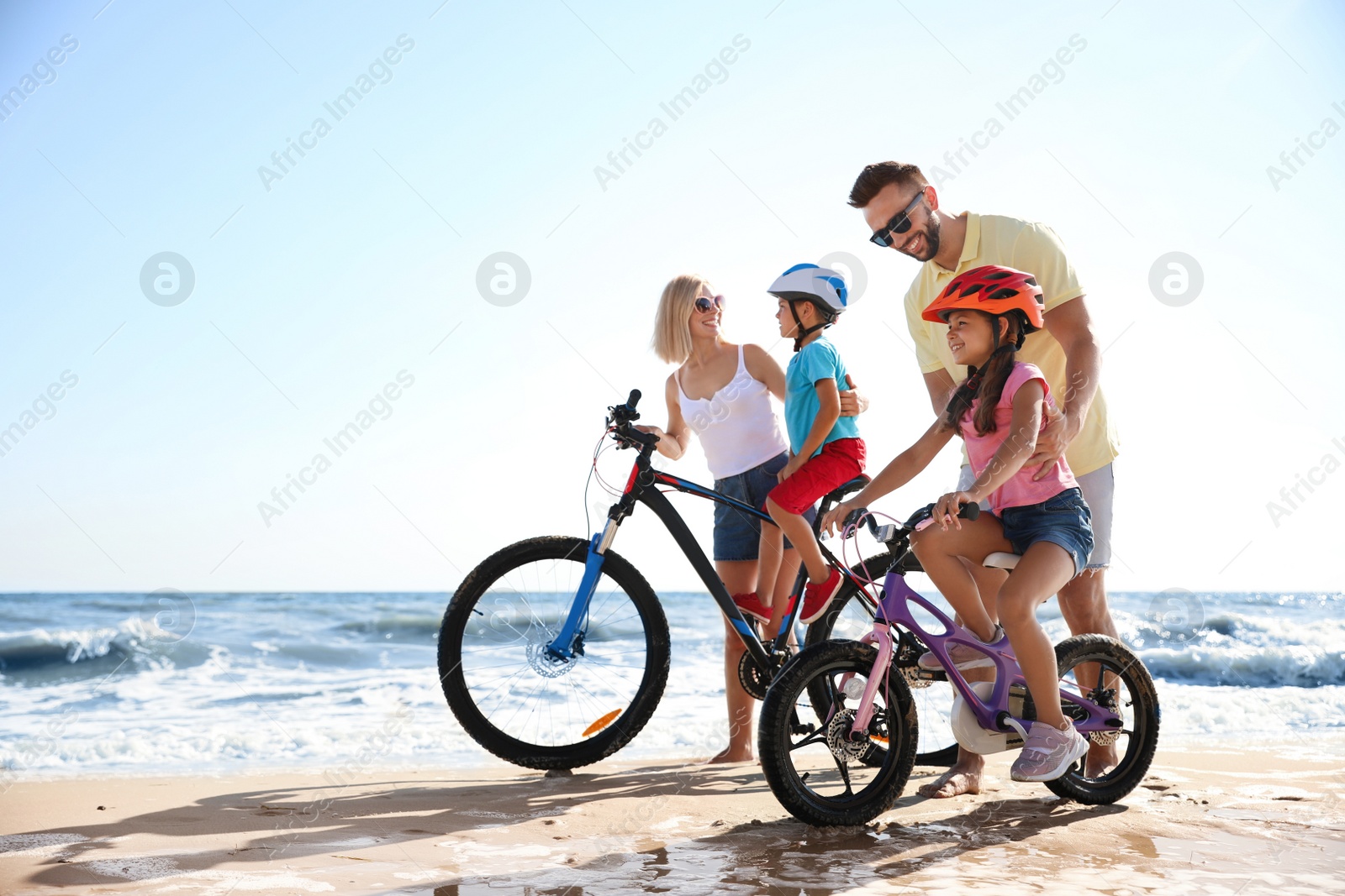 Photo of Happy parents teaching children to ride bicycles on sandy beach near sea