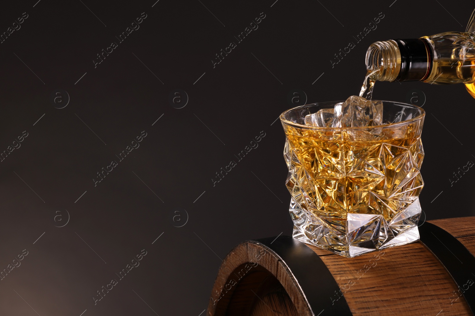 Photo of Pouring whiskey from bottle into glass on wooden barrel against dark background, closeup. Space for text