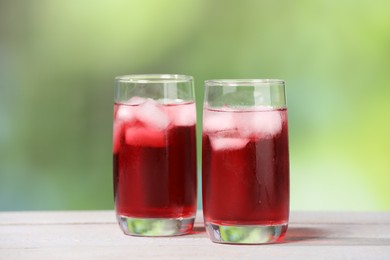 Refreshing hibiscus tea with ice cubes in glasses on white wooden table against blurred green background