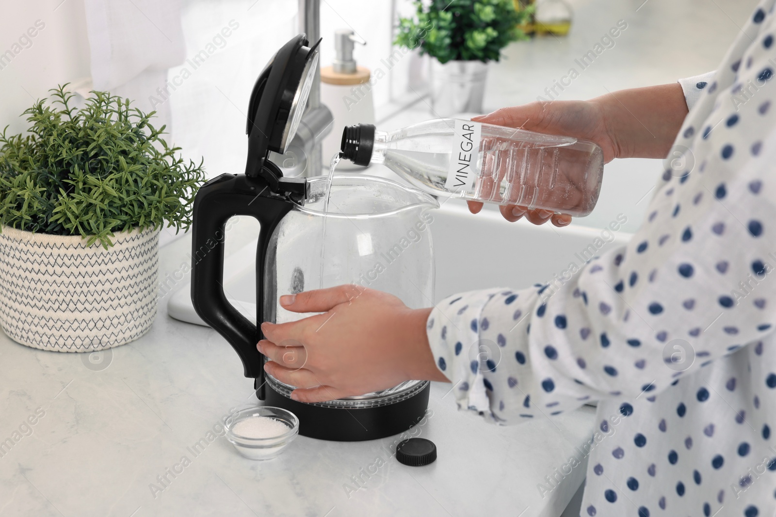 Photo of Woman pouring vinegar from bottle into electric kettle in kitchen, closeup