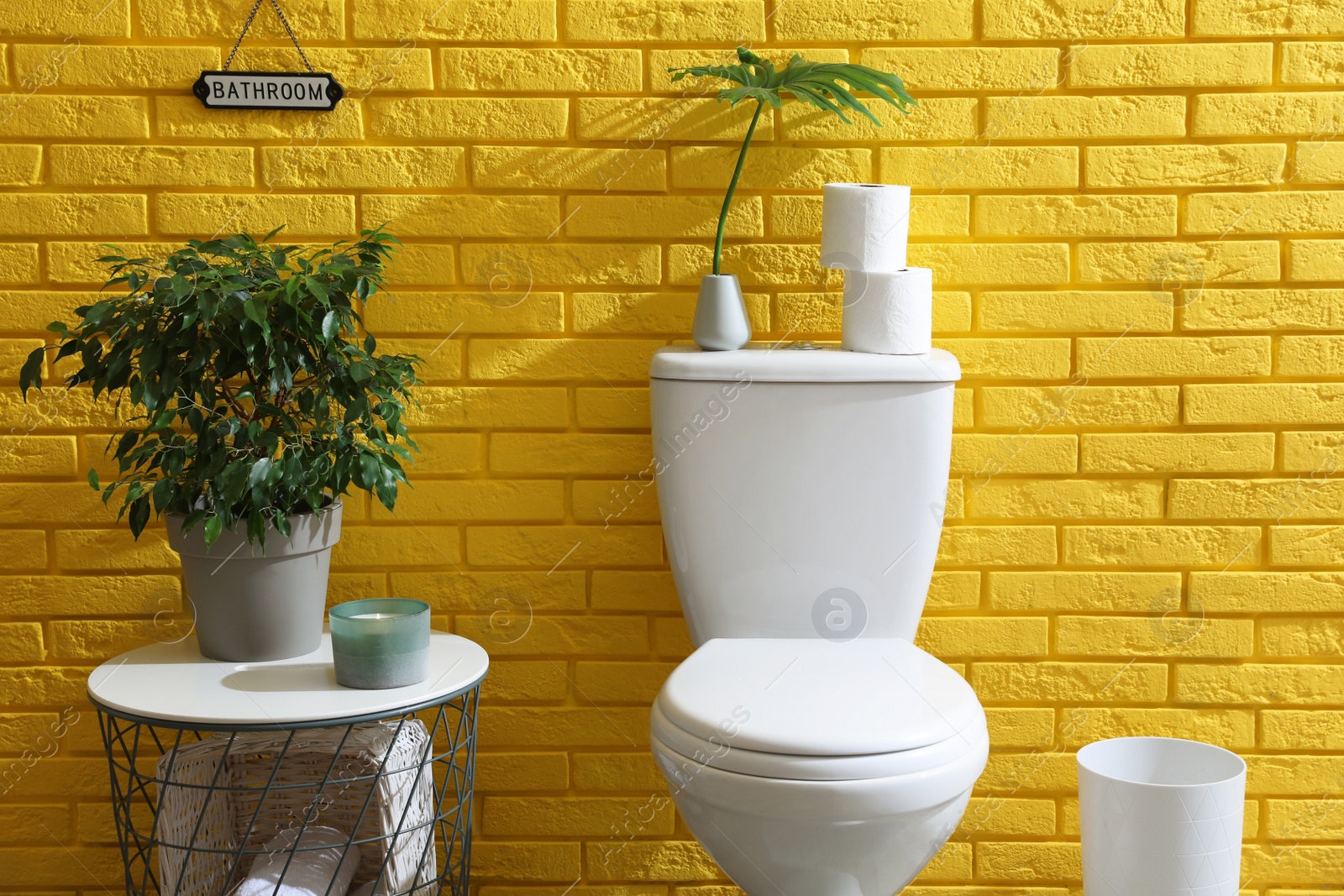 Photo of Stylish bathroom with toilet bowl, green plants and decor elements near yellow brick wall. Interior design