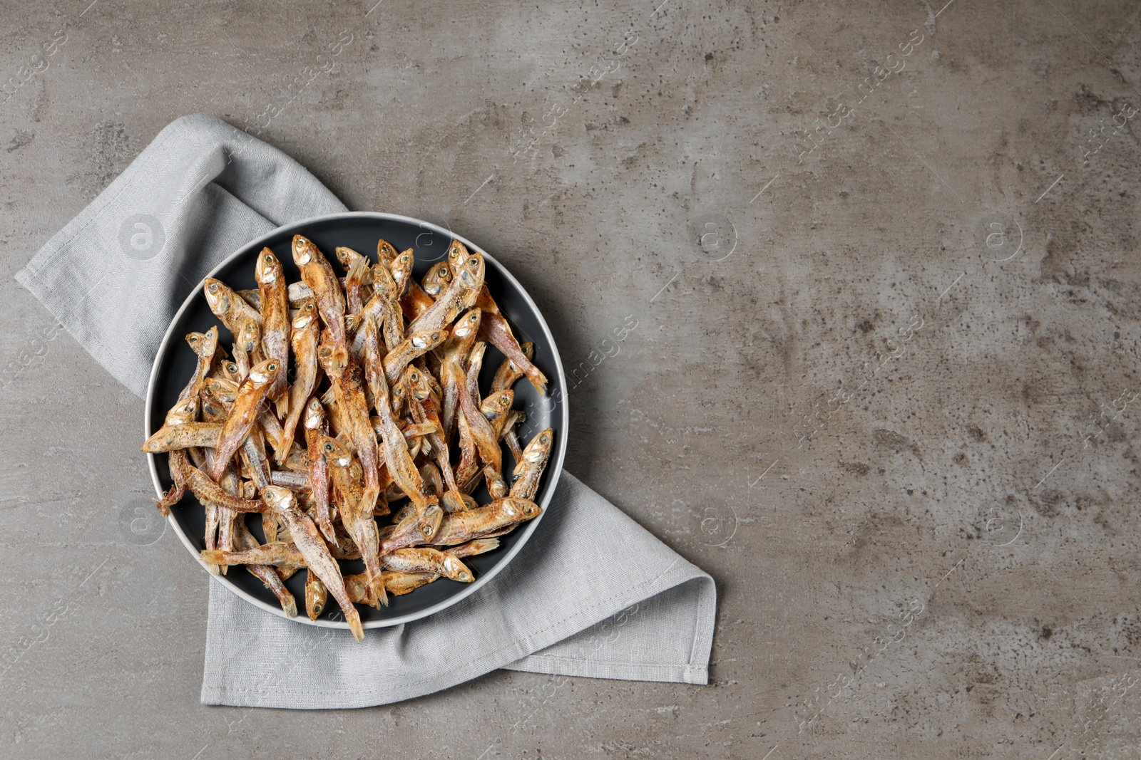 Photo of Plate of tasty dried anchovies and napkin on grey table, top view. Space for text