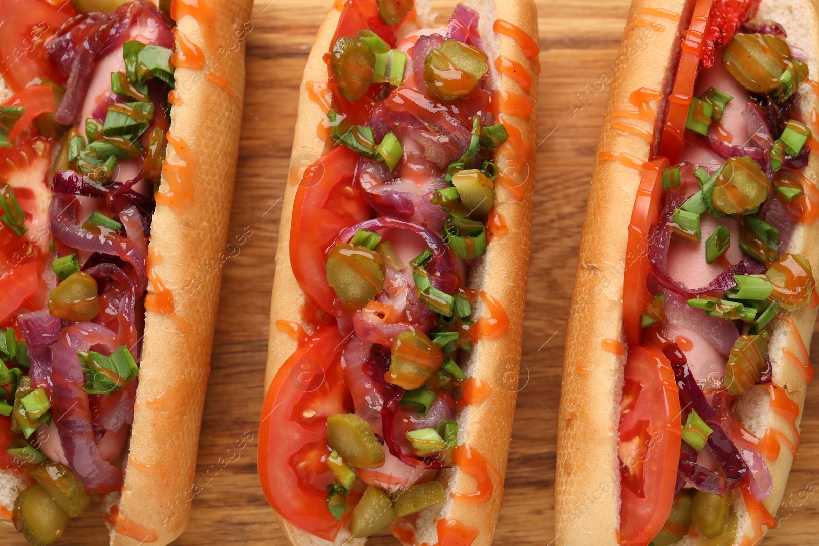 Photo of Tasty hot dogs with green onion, tomato, pickles and sauce on wooden table, above view