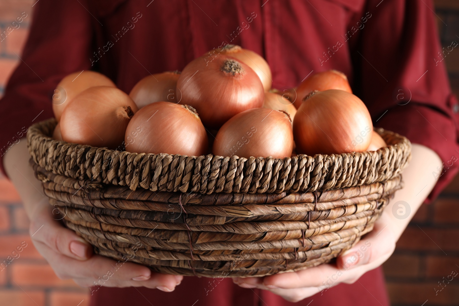 Photo of Woman holding wicker basket with ripe onions against red brick wall, closeup