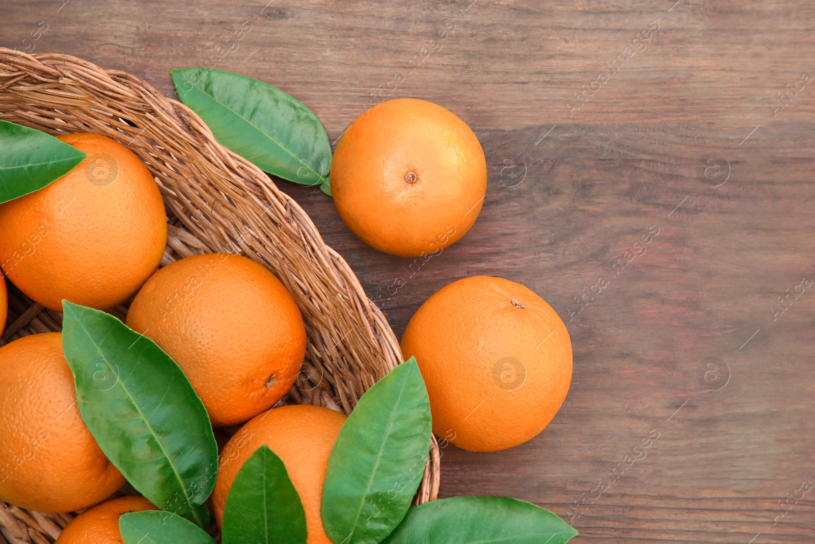 Photo of Wicker basket, ripe juicy oranges and green leaves on wooden table, flat lay. Space for text