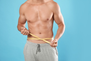 Young man with slim body using measuring tape on light blue background, closeup view