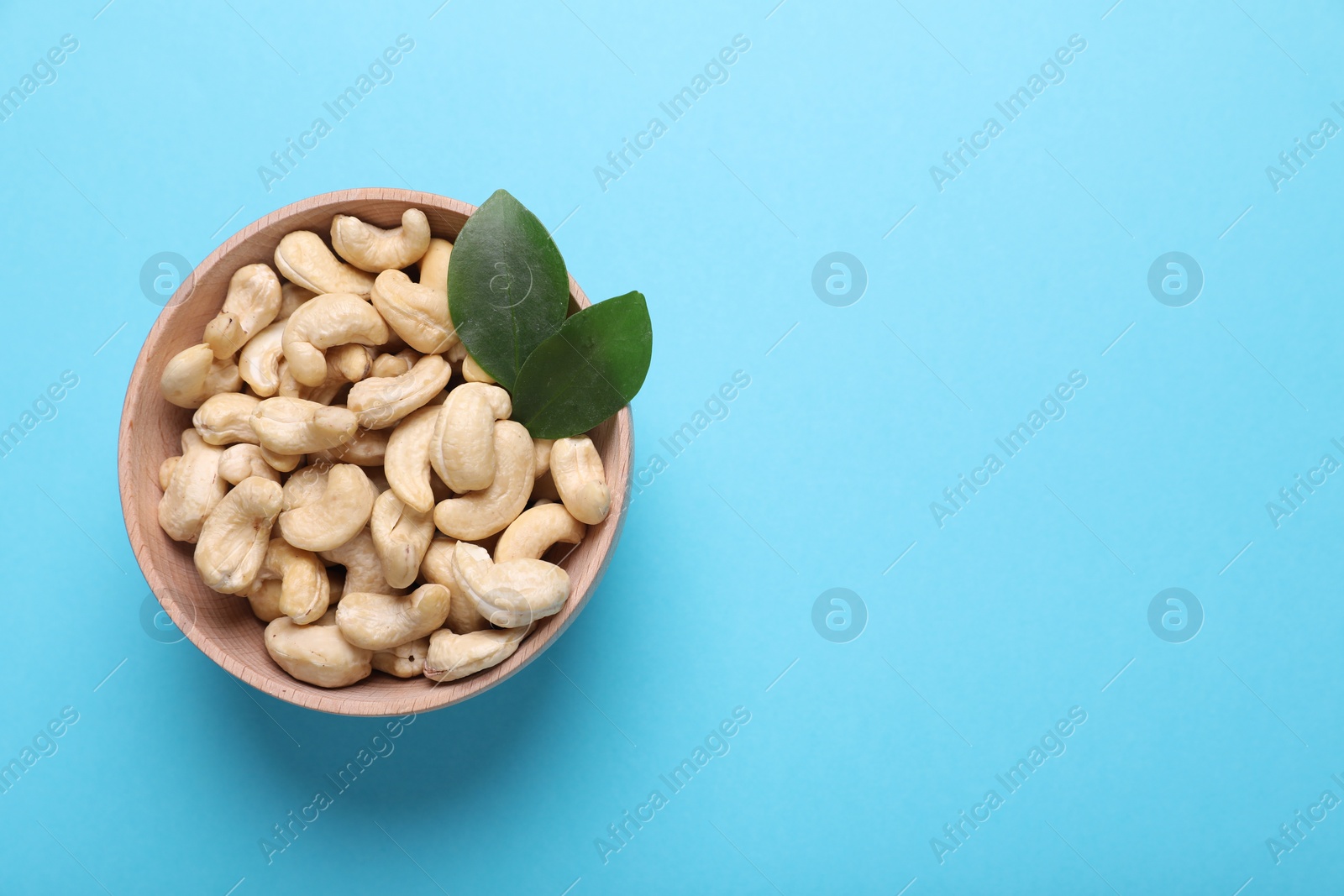 Photo of Tasty cashew nuts and green leaves in bowl on light blue background, top view. Space for text