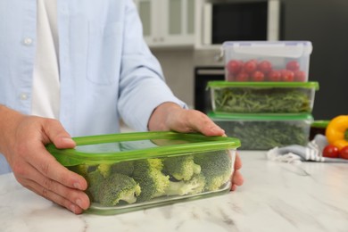 Man holding glass container with fresh broccoli at white marble table in kitchen, closeup. Food storage