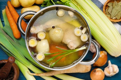 Pot with tasty bouillon and different ingredients on blue wooden table, flat lay