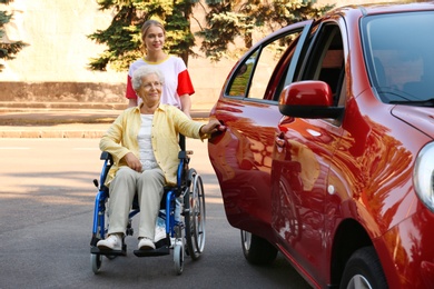 Photo of Young woman helping disabled grandmother in wheelchair to get into car outdoors