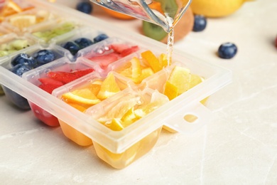 Pouring water into ice cube tray with different fruits and berries on table, closeup