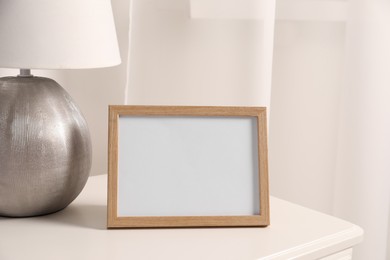 Empty square frame and lamp on white bedside table in room