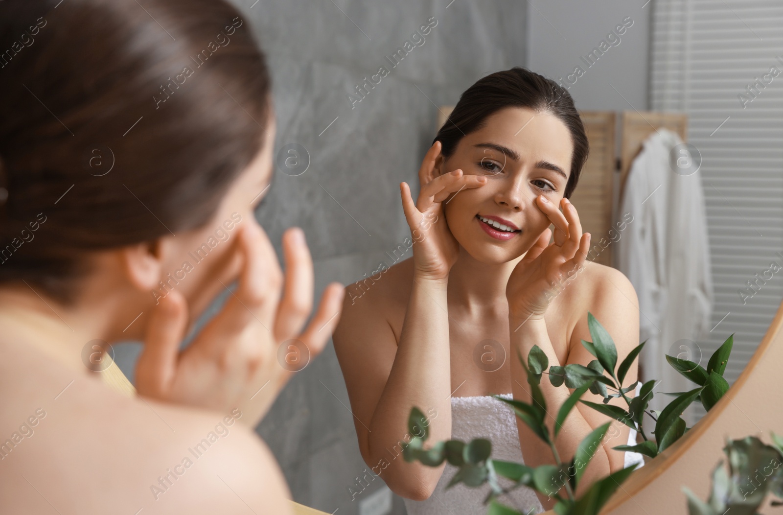 Photo of Young woman massaging her face near mirror in bathroom