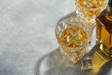 Photo of Whiskey with ice cubes in glasses and bottle on grey textured table, space for text