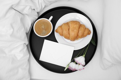 Tray with tasty croissant, cup of coffee and flowers on white bed, top view