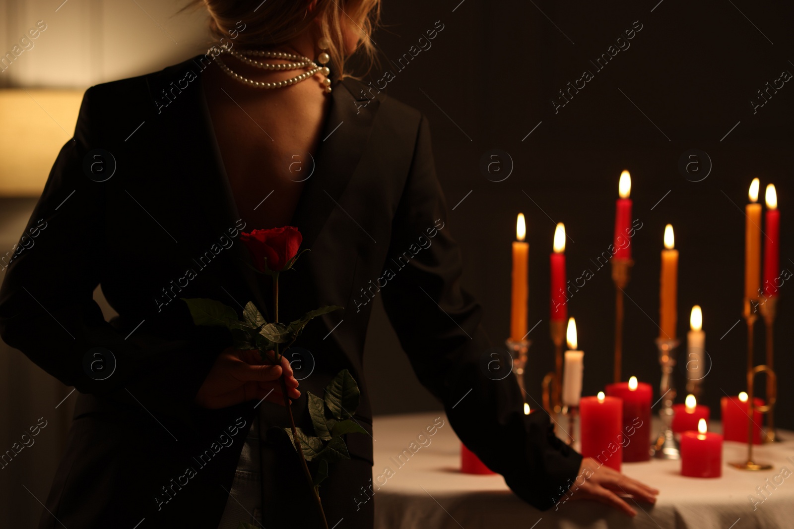 Photo of Woman holding beautiful rose near table with burning candles at night, back view