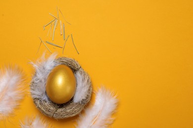 Photo of Shiny golden egg with feathers in nest on orange background, flat lay. Space for text