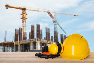 Image of Safety equipment. Hard hat, gloves and protective headphones on wooden surface near construction site with tower crane outdoors, space for text