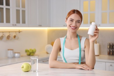 Photo of Happy young woman with bottle of pills at table in kitchen, space for text. Weight loss