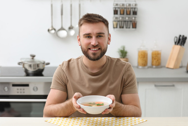 Photo of Young man holding bowl of tasty soup at table in kitchen
