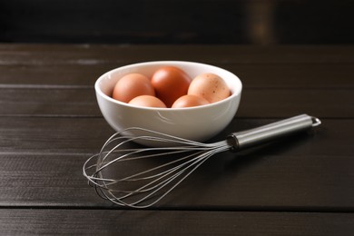 Metal whisk and raw eggs in bowl on dark wooden table, closeup