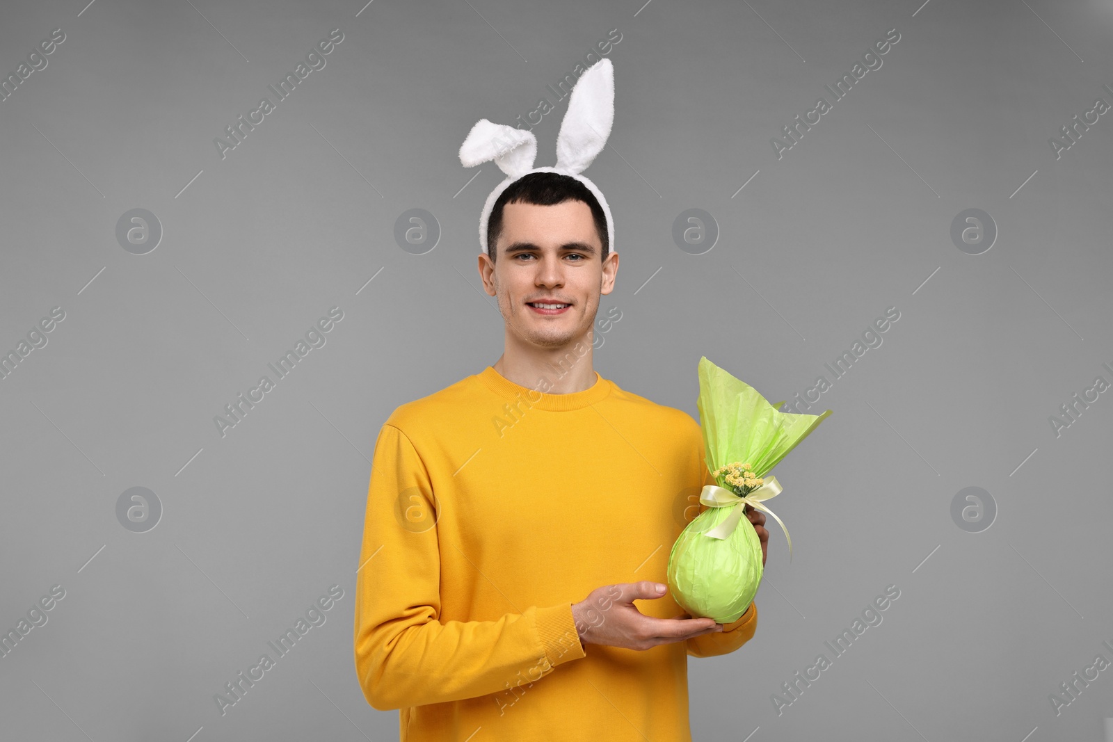 Photo of Easter celebration. Handsome young man with bunny ears holding wrapped gift on grey background