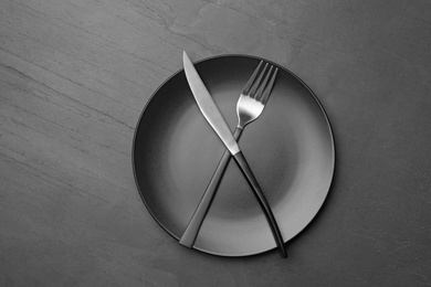 Stylish ceramic plate with crossed fork and knife on dark background, top view