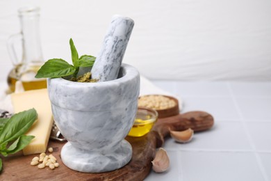 Photo of Mortar with pestle and different ingredients for cooking tasty pesto sauce on white tiled table. Space for text