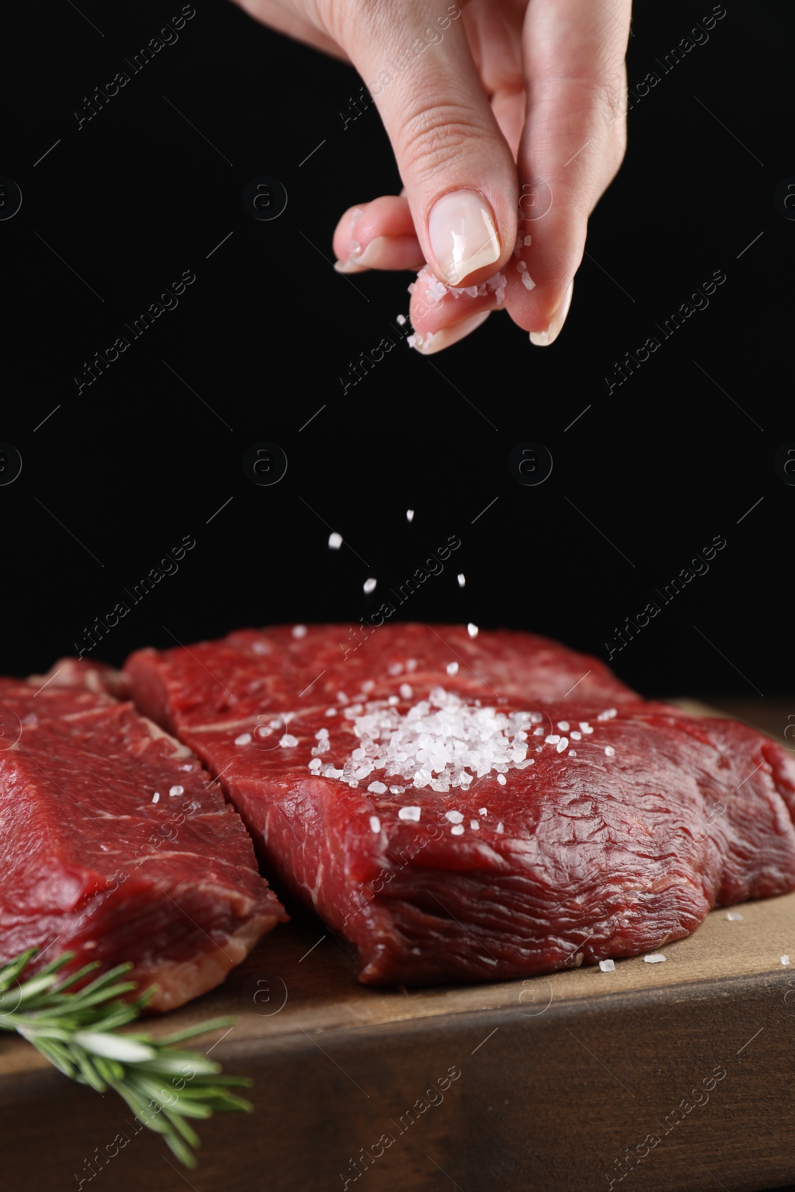 Photo of Woman salting fresh raw beef steak at table against black background, closeup