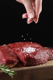 Woman salting fresh raw beef steak at table against black background, closeup