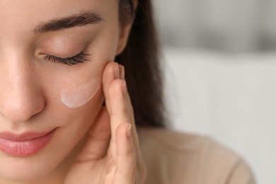 Photo of Young woman with dry skin applying cream onto her face against blurred background, closeup. Space for text