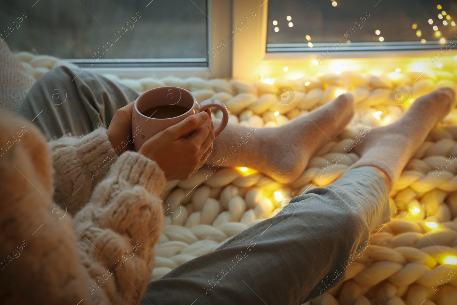 Photo of Woman with drink sitting on soft plaid near window in evening, closeup