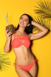 Beautiful young woman in stylish bikini with cocktail and tropical leaves on yellow background