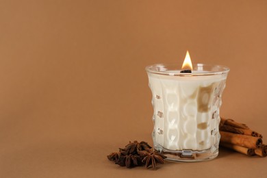 Photo of Burning soy candle, anise stars and cinnamon sticks on brown background, space for text