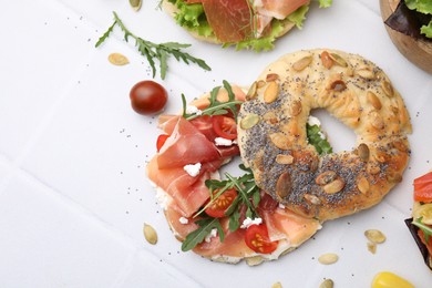 Photo of Tasty bagel with cured ham, cheese, tomatoes and arugula on white tiled table, top view. Space for text