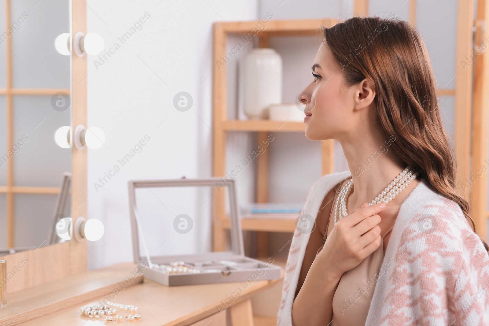 Photo of Young woman trying on elegant pearl necklace at dressing table indoors, space for text