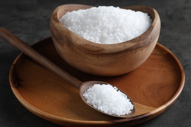 Natural salt in wooden bowl and spoon on dark grey table, closeup