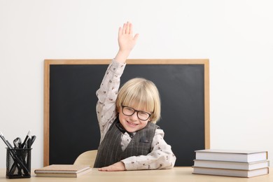 Photo of Happy little school child raising hand while sitting at desk with books near chalkboard in classroom