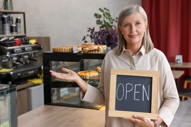 Photo of Smiling business owner holding open sign and inviting to come into her cafe, space for text