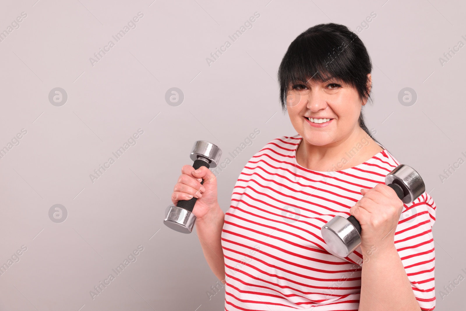 Photo of Happy overweight mature woman doing exercise with dumbbells on grey background, space for text