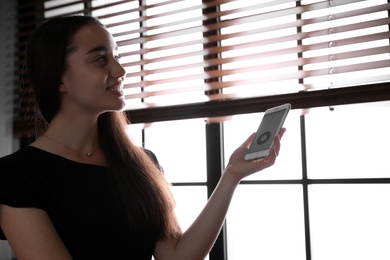 Photo of Woman using smart home application on phone to control window blinds indoors
