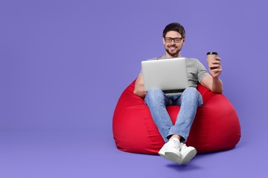 Happy man with laptop and coffee sitting in beanbag chair against lilac background, space for text