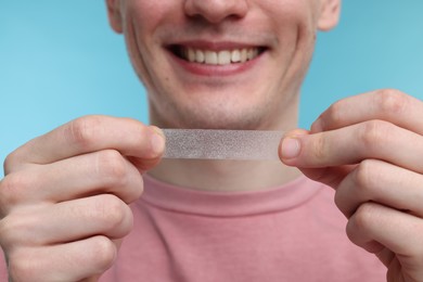 Young man with whitening strip on light blue background, closeup