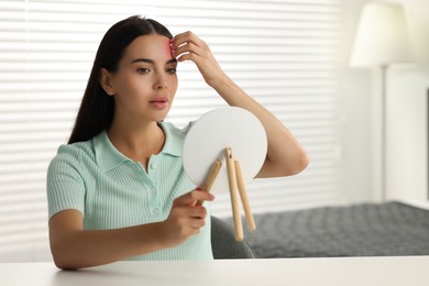 Photo of Suffering from allergy. Young woman looking in mirror and scratching her forehead at white table indoors, space for text