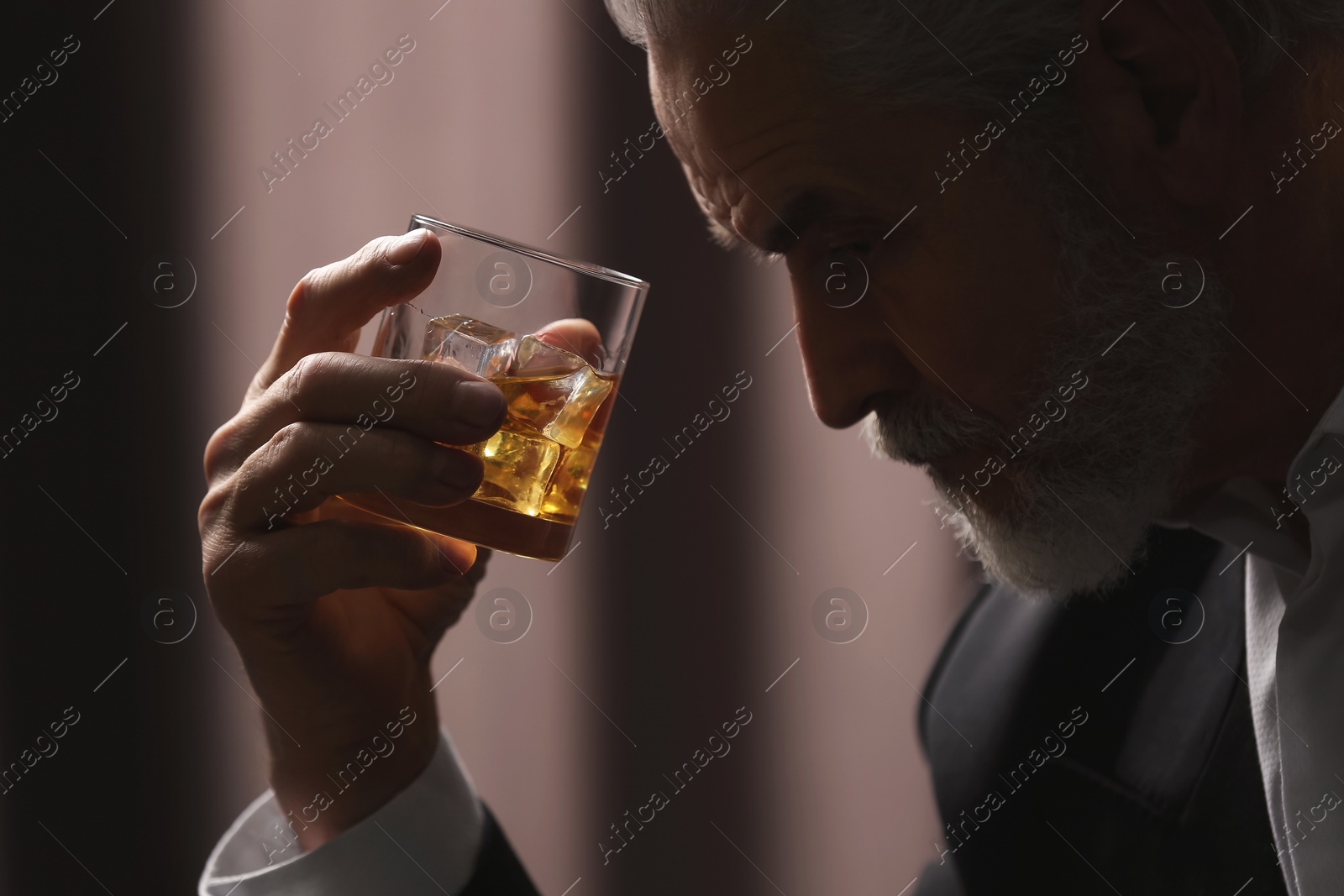 Photo of Senior man holding glass of whiskey with ice cubes on brown background, closeup